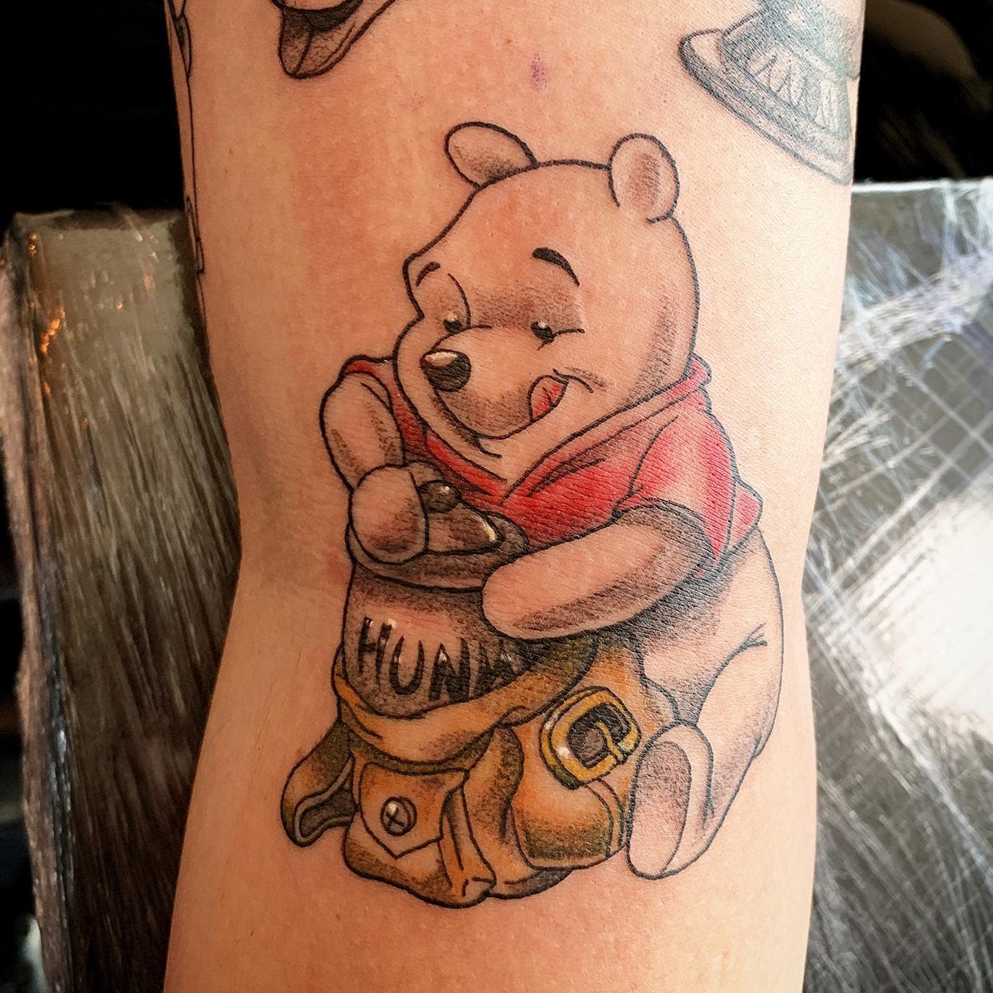 Tattoo of Winnie the Pooh happily holding a honey pot labeled 'Hunny' on a person's arm